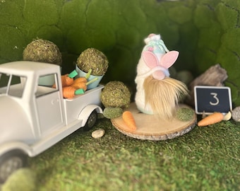 Puede incluir: Un camión de juguete blanco transporta zanahorias naranjas con tapas verdes y bolas de musgo. Un gnomo con barba blanca, orejas de conejo y un sombrero está sentado en una rodaja de madera. Una pequeña pizarra muestra el número 3.