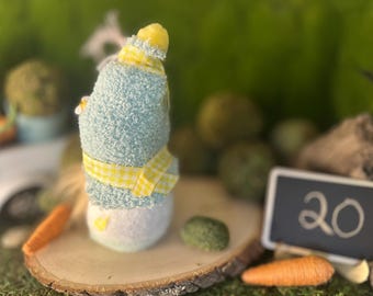 Puede incluir: Figura de conejito de Pascua hecha a mano con cuerpo azul claro, detalles amarillos y parte inferior blanca. El conejito está decorado con una cinta a cuadros amarilla y blanca. Una pequeña pizarra con el número 20 está en el fondo.