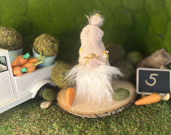 Puede incluir: Un gnomo de Pascua caprichoso con un sombrero rosa y barba blanca, adornado con detalles de abeja, está sentado sobre una rodaja de madera. Zanahorias en miniatura y un camión blanco lleno de zanahorias están cerca, junto a una pequeña pizarra con el número 5.