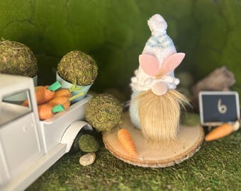 Puede incluir: Una composición temática de Pascua con un camión de juguete blanco que transporta zanahorias naranjas y bolas de musgo. Un gnomo con sombrero blanco, orejas de conejo rosas y larga barba beige está sobre una rodaja de madera. Una pequeña pizarra muestra el número 6.