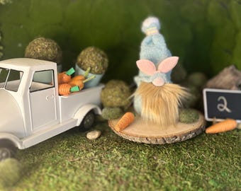 Puede incluir: Un camión de juguete blanco transporta zanahorias, junto a un gnomo con orejas de conejo y una larga barba. Una pequeña pizarra muestra el número 2. La escena está sobre musgo verde.