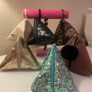 May include: A collection of triangular cosmetic bags in various colors and textures. The bags include a gold sequin, black woven, leopard print, and a teal sequin bag. Each bag has a zipper closure.