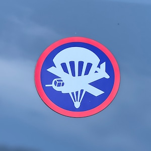 Può includere: Un adesivo rotondo con sfondo blu e una grafica bianca di un paracadute e un aereo. L'adesivo ha un bordo rosso. L'immagine è ambientata contro un cielo nuvoloso.