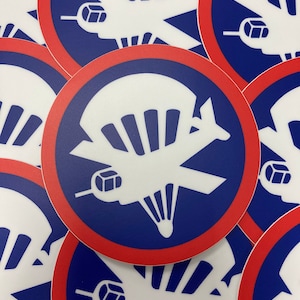 May include: Close-up of a stack of round stickers with a white silhouette of a plane with a parachute on a blue background with a red border.