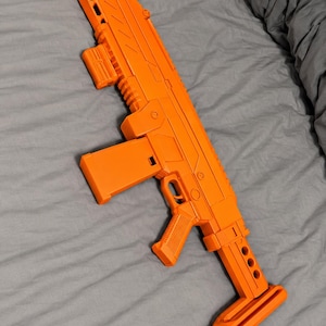 May include: A bright orange toy rifle featuring a rectangular magazine, a textured pistol grip, a trigger guard, and a stock with three circular cutouts. The body of the toy rifle has a modular design with visible vents along the barrel and a top rail.