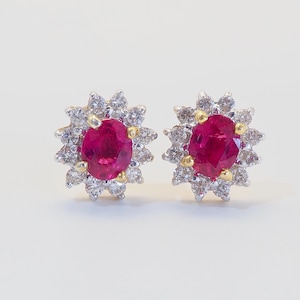 May include: A pair of stud earrings featuring a vibrant red oval gemstone surrounded by a halo of smaller, clear gemstones. The earrings have a gold and silver setting, creating a striking contrast. The design is reminiscent of a flower.