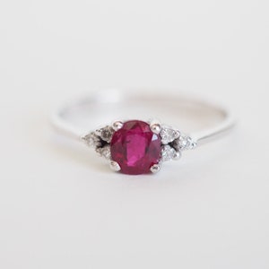 May include: A silver ring with a central oval ruby, complemented by smaller, clear accent stones. The ruby is a deep red hue. The ring's band is slim and polished, offering a classic design. The ring is set against a plain white backdrop, highlighting the gemstones.