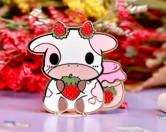 Puede incluir: Un pin de esmalte de vaca blanca con mejillas rosadas y una fresa en la boca. La vaca lleva una fresa roja en cada oreja. El pin está sobre una superficie morada con un ramo de flores en el fondo.