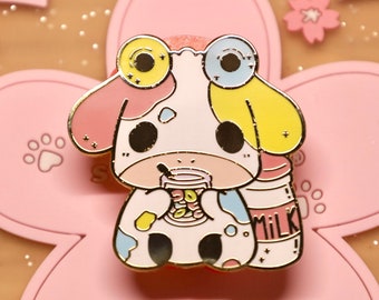Puede incluir: Un pin de esmalte dorado con una vaca de dibujos animados con salpicaduras de pintura de colores, que lleva gafas y sostiene una taza de leche. La vaca lleva una diadema amarilla y rosa y tiene un fondo blanco con un patrón de flores rosas y blancas.