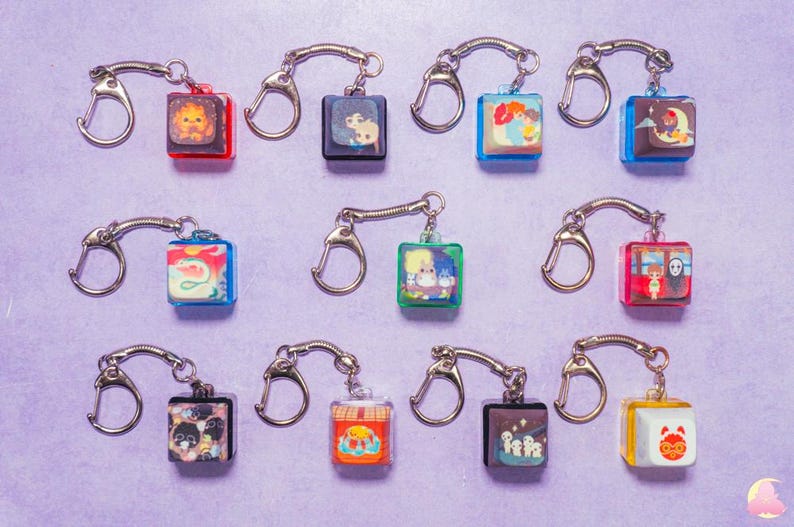 May include: Twelve colorful keychains with different cartoon characters and designs. Each keychain features a square charm with a clear plastic cover and a silver metal key ring.