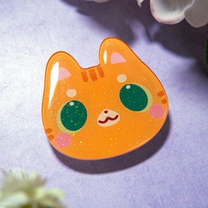 May include: Orange cat-shaped resin charm with glitter accents. The charm features a cute cat face with green eyes, pink cheeks, and a small, smiling mouth. The background is a soft purple with white flowers.
