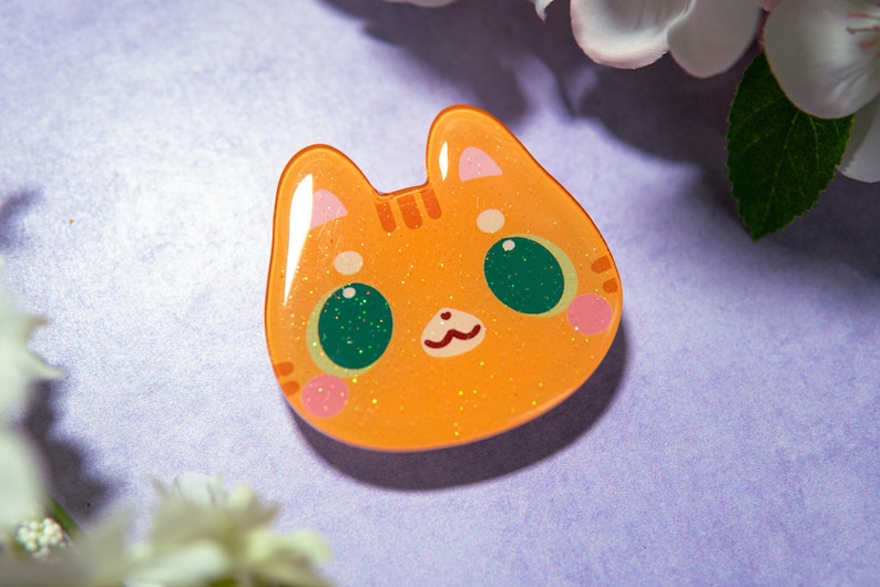 May include: Orange cat-shaped resin charm with glitter accents. The charm features a cute cat face with green eyes, pink cheeks, and a small, smiling mouth. The background is a soft purple with white flowers.