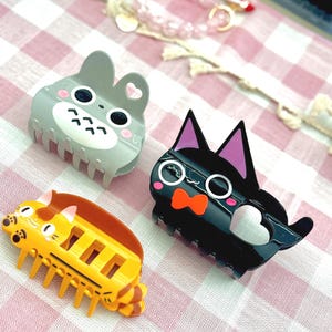 May include: Three novelty hair clips in the shape of cartoon animals. One is grey with a bunny design, one is black with a cat design, and the third is yellow with a cat design. The clips are on a pink and white checked surface.