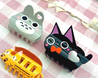 May include: Three decorative hair clips in the shape of cartoon animals. One is gray with a bunny design, one is black with a cat design, and the third is yellow with a cat design. The clips are on a pink and white checkered surface.