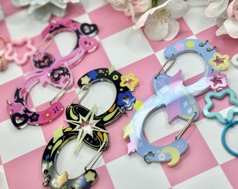 Puede incluir: Clips de mosquetón decorativos en varios diseños. Un clip es rosa con detalles florales y de corazón, otro es negro con un diseño de estrella y un tercero es azul claro con estrellas y lunas. Cada clip tiene un cierre plateado.