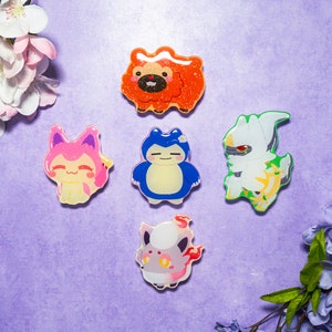 May include: Five colorful cartoon character pins on a purple background. The pins are of different sizes and shapes, and each features a different character. The characters are all smiling and have large eyes. The pins are surrounded by pink and purple flowers.