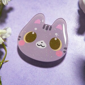 May include: A lavender cat-shaped pin with glitter accents. The cat has large brown eyes, pink cheeks, and small pink ears. The pin is set against a purple background with white flowers.