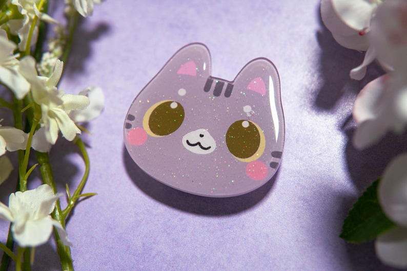 May include: A lavender cat-shaped pin with glitter accents. The cat has large brown eyes, pink cheeks, and small pink ears. The pin is set against a purple background with white flowers.
