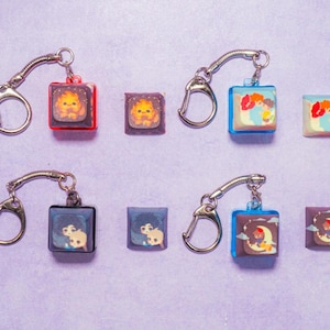 May include: Six keychains with different colorful square charms featuring cartoon characters. Each keychain has a silver metal ring and a silver chain.