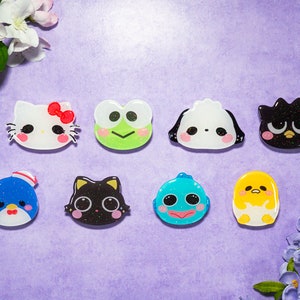 Może przedstawiać: Osiem kolorowych figurek z żywicy z postaciami z kreskówek, w tym Hello Kitty, Keropi, Pochacco, Kero Kero Keroppi, Gudetama i pingwin. Figurki znajdują się na fioletowym tle.
