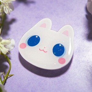 May include: A white cat-shaped pin with glitter accents, featuring large blue eyes with glitter, pink cheeks, and a small pink mouth. The pin is set against a purple background with white flowers.