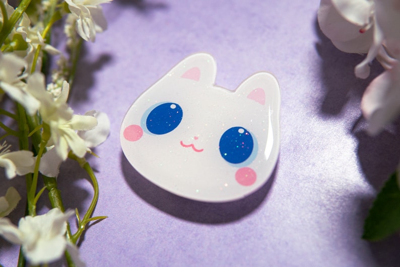 May include: A white cat-shaped pin with glitter accents, featuring large blue eyes with glitter, pink cheeks, and a small pink mouth. The pin is set against a purple background with white flowers.