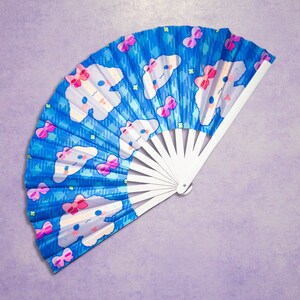 May include: A blue and white hand fan with a repeating pattern of white clouds with pink bows and a cartoon character's face. The fan is folded and has a white handle.