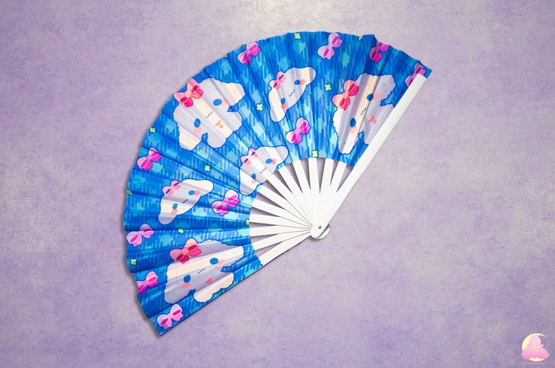 May include: A blue and white hand fan with a repeating pattern of white clouds with pink bows and a cartoon character's face. The fan is folded and has a white handle.