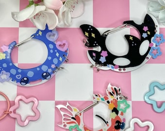 May include: Three decorative carabiner clips in the image. One is blue with hearts and dots, another is black and white with star accents, and the third is colorful with floral and star designs. Pink and white star-shaped keychains are also visible.