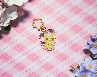May include: A clear acrylic keychain featuring a yellow cartoon character with a pink background. The character has a surprised expression and is holding a small flower. The keychain is attached to a gold metal chain with a flower charm.