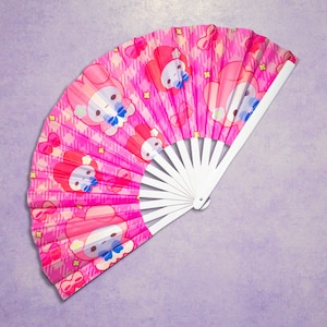 May include: A pink hand fan with a white handle. The fan features a repeating pattern of a cartoon character with a pink bow and a white background.