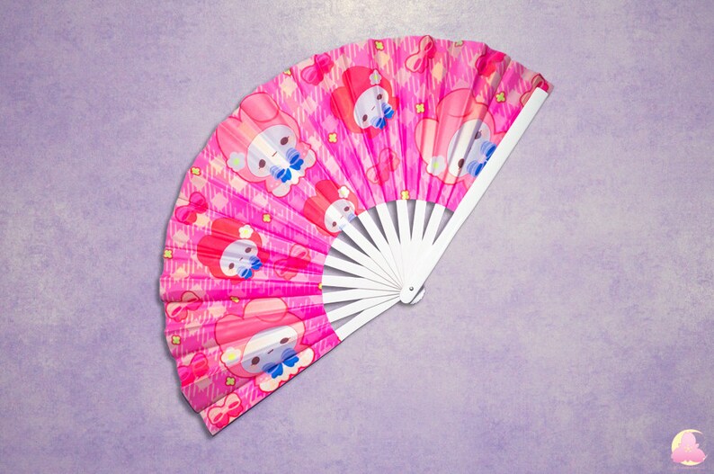 May include: A pink hand fan with a white handle. The fan features a repeating pattern of a cartoon character with a pink bow and a white background.