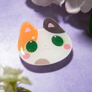 May include: A cat-shaped resin pin with a white base, orange and black ears, and green glittery eyes. The pin has pink cheeks and a small, smiling mouth. The background is a soft purple with white flowers.