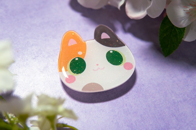 May include: A cat-shaped resin pin with a white base, orange and black ears, and green glittery eyes. The pin has pink cheeks and a small, smiling mouth. The background is a soft purple with white flowers.