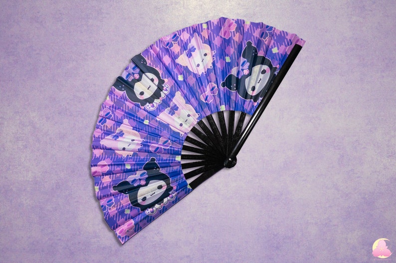 May include: A purple and pink hand fan with a black handle. The fan features a repeating pattern of cartoon characters with black eyes and white bows.