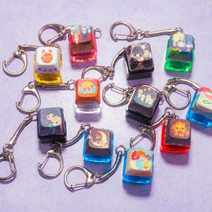 May include: A collection of 12 colorful keychains featuring various cartoon characters and designs. Each keychain is made of a clear plastic material with a metal ring and a metal chain. The keychains are arranged on a purple surface.