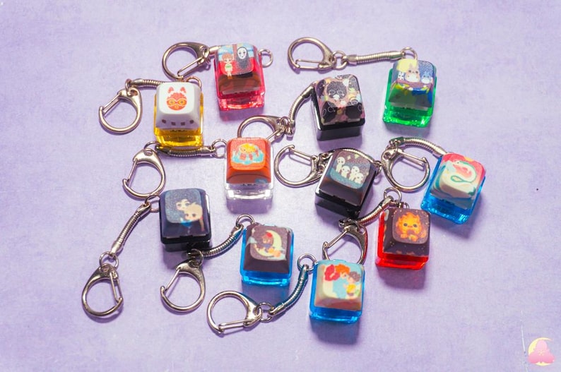 May include: A collection of 12 colorful keychains featuring various cartoon characters and designs. Each keychain is made of a clear plastic material with a metal ring and a metal chain. The keychains are arranged on a purple surface.