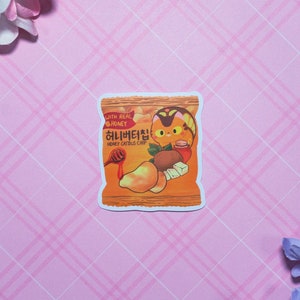 以下が含まれることがあります： 韓国語と英語で「Honey Catbus Chip」と「With Real Honey」と書かれた、漫画のハチミツ猫チップ袋のステッカー。袋はオレンジとイエローで、漫画の猫がハチミツの壺を持っています。