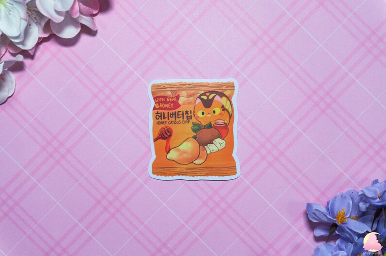 以下が含まれることがあります： 韓国語と英語で「Honey Catbus Chip」と「With Real Honey」と書かれた、漫画のハチミツ猫チップ袋のステッカー。袋はオレンジとイエローで、漫画の猫がハチミツの壺を持っています。