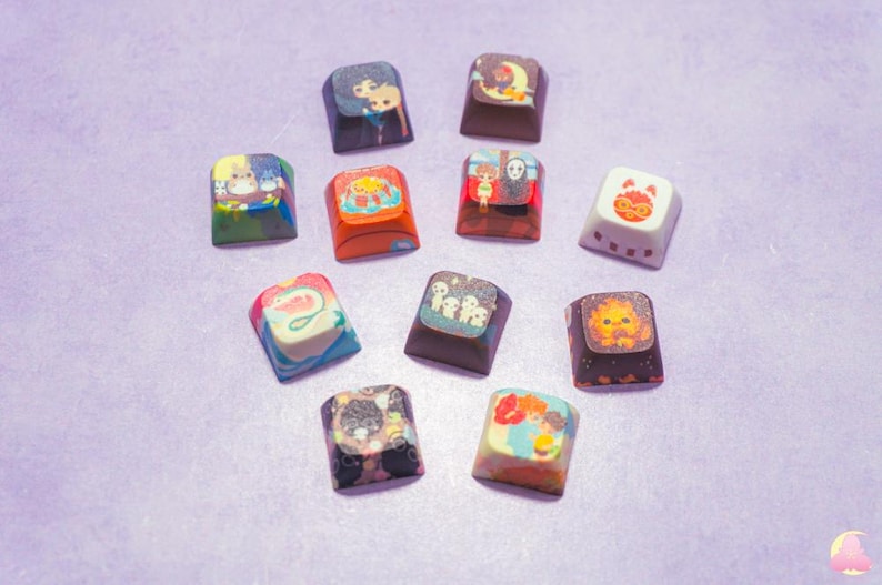 May include: Twelve colorful square keycaps with various cartoon characters and designs. The keycaps are arranged in a semi-circle on a purple surface.