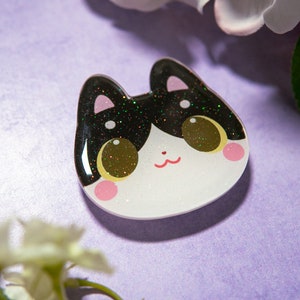 May include: A cat-shaped resin pin with a black and glittery top, white face, and pink cheeks. The cat has large, golden eyes and a small red mouth. The pin is set against a purple background with white flowers.