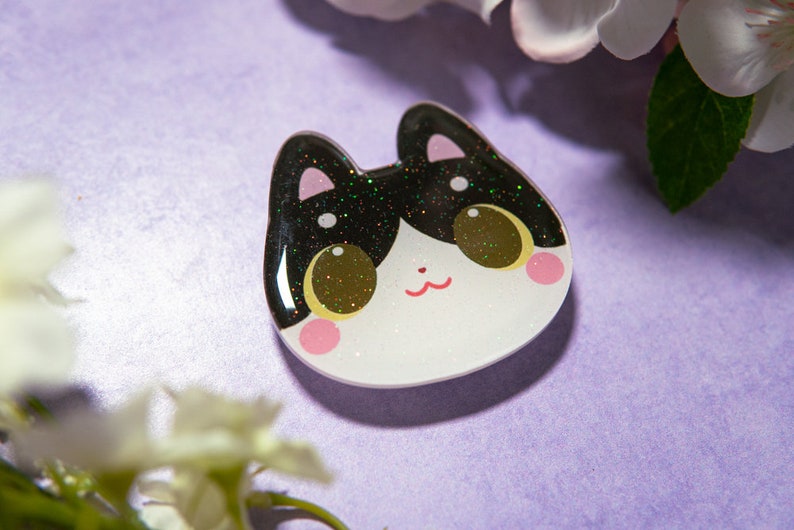 May include: A cat-shaped resin pin with a black and glittery top, white face, and pink cheeks. The cat has large, golden eyes and a small red mouth. The pin is set against a purple background with white flowers.