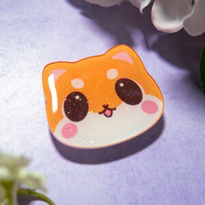 May include: A cute, orange Shiba Inu face-shaped pin with glitter accents. The pin has a white base, pink cheeks, and brown eyes. The background is a light purple color with white flowers.