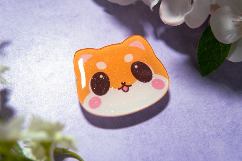 May include: A cute, orange Shiba Inu face-shaped pin with glitter accents. The pin has a white base, pink cheeks, and brown eyes. The background is a light purple color with white flowers.