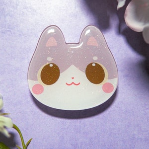May include: A cat-shaped resin charm with a glittery, light purple and white design. The charm features large brown eyes, pink cheeks, and a small red mouth. The cat's ears are also pink.