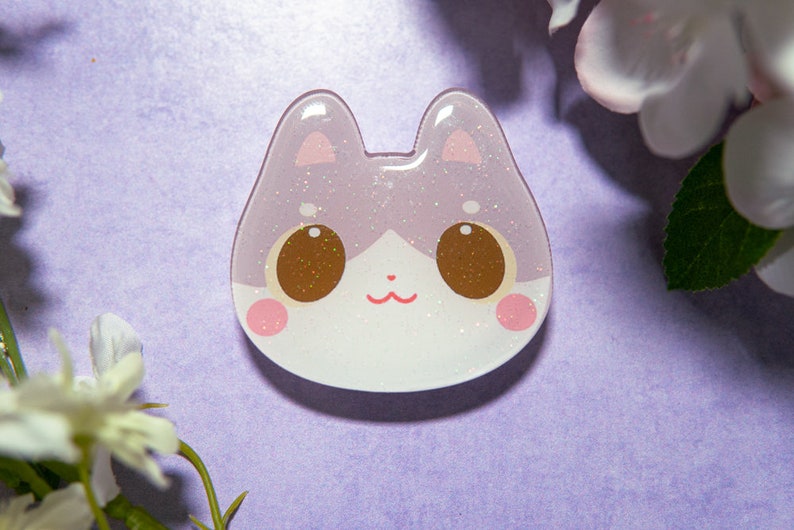 May include: A cat-shaped resin charm with a glittery, light purple and white design. The charm features large brown eyes, pink cheeks, and a small red mouth. The cat's ears are also pink.