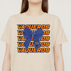 May include: Beige t-shirt featuring the word "VAQUEROS" in orange and yellow block letters, with a blue bow overlay. A cartoon cowboy is in the center of the design. The shirt has short sleeves and a crew neck.