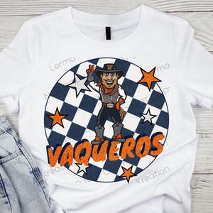 May include: White t-shirt featuring a cartoon cowboy character with a black hat, orange scarf, and vest. The design includes the word "VAQUEROS" in orange glitter lettering, surrounded by stars and a checkered background.
