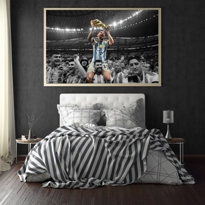 May include: A framed print of a soccer player holding a trophy, surrounded by teammates, in a stadium setting. The image is mostly black and white, with the player's jersey in blue and white stripes. The print hangs above a bed with a striped blanket.