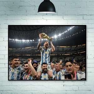 May include: A framed print of a soccer team celebrating a victory. The player in the center, wearing a blue and white striped jersey, holds a trophy aloft. Teammates surround him, some on shoulders, in a stadium setting.